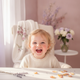 Portrait of a smiling toddler, around 2-5 years old, with a happy expression. The background is a soft blur of dusty rose and